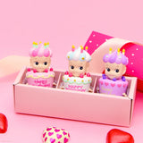 Three vibrant, doll-like figures from the Sonny Angel Happy Birthday Cake Gift series are nestled in a pink blind box. Red and pink heart shapes are scattered in the foreground, adding an extra touch of charm.