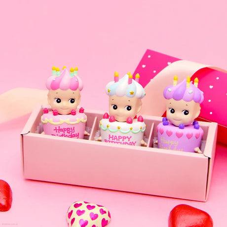 Three vibrant, doll-like figures from the Sonny Angel Happy Birthday Cake Gift series are nestled in a pink blind box. Red and pink heart shapes are scattered in the foreground, adding an extra touch of charm.