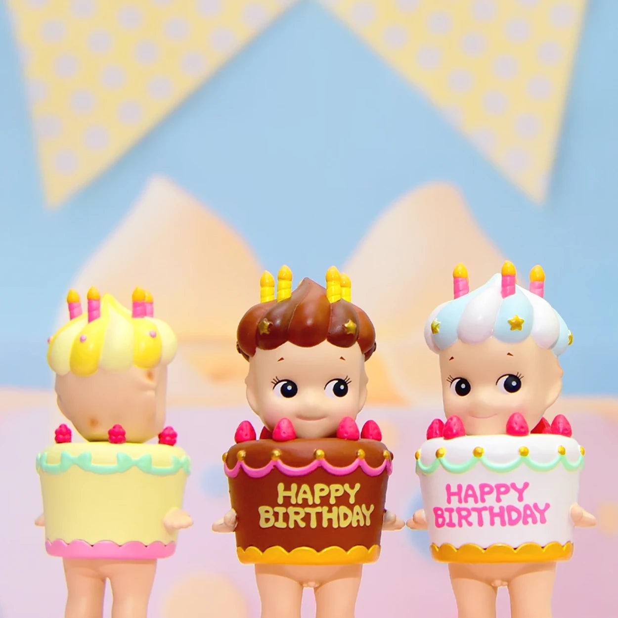 The Sonny Angel Happy Birthday Cake Gift features three adorable figures from the Birthday Cake Series, each dressed in a "Happy Birthday" cake outfit adorned with colorful candles and decorations, set against a pastel backdrop with festive bunting. These charming Sonny Angels add joy to any celebration.