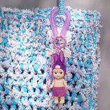 A Sonny Angel from the Candy Store Series, wearing a purple bunny hood and outfit, hangs from a purple clip against a textured blue and white knitted background.