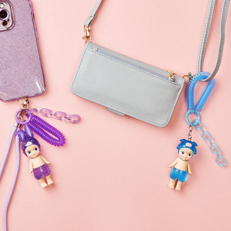 On a pastel pink background, a purple smartphone is displayed alongside two Sonny Angel Candy Store Series toy figurines from the Blind Box on keychains and a light blue purse with straps.