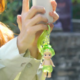 Someone holds a smartphone next to a green keychain with a tiny doll in a frog hat and shorts from the Sonny Angel Candy Store Series - Blind Box by Sonny Angel.