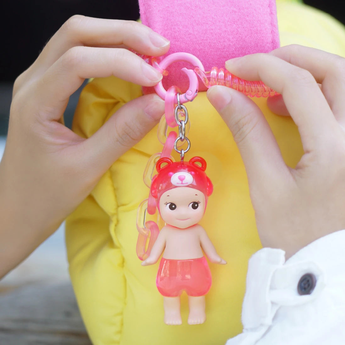 Hands holding a pink keychain attached to a yellow bag, featuring a small doll in a red bear hat and shorts, echoing the whimsical charm of Sonny Angel Candy Store Series - Blind Box by Sonny Angel.