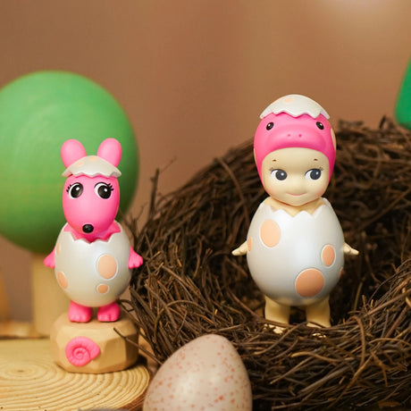 Two egg-themed toys from the Sonny Angel Dinosaur Series by Sonny Angel: one featuring a pink animal and another as a figure in a nest on a wooden surface, accompanied by a green abstract tree. A speckled egg nearby enhances the charm of these limited edition prehistoric pals.