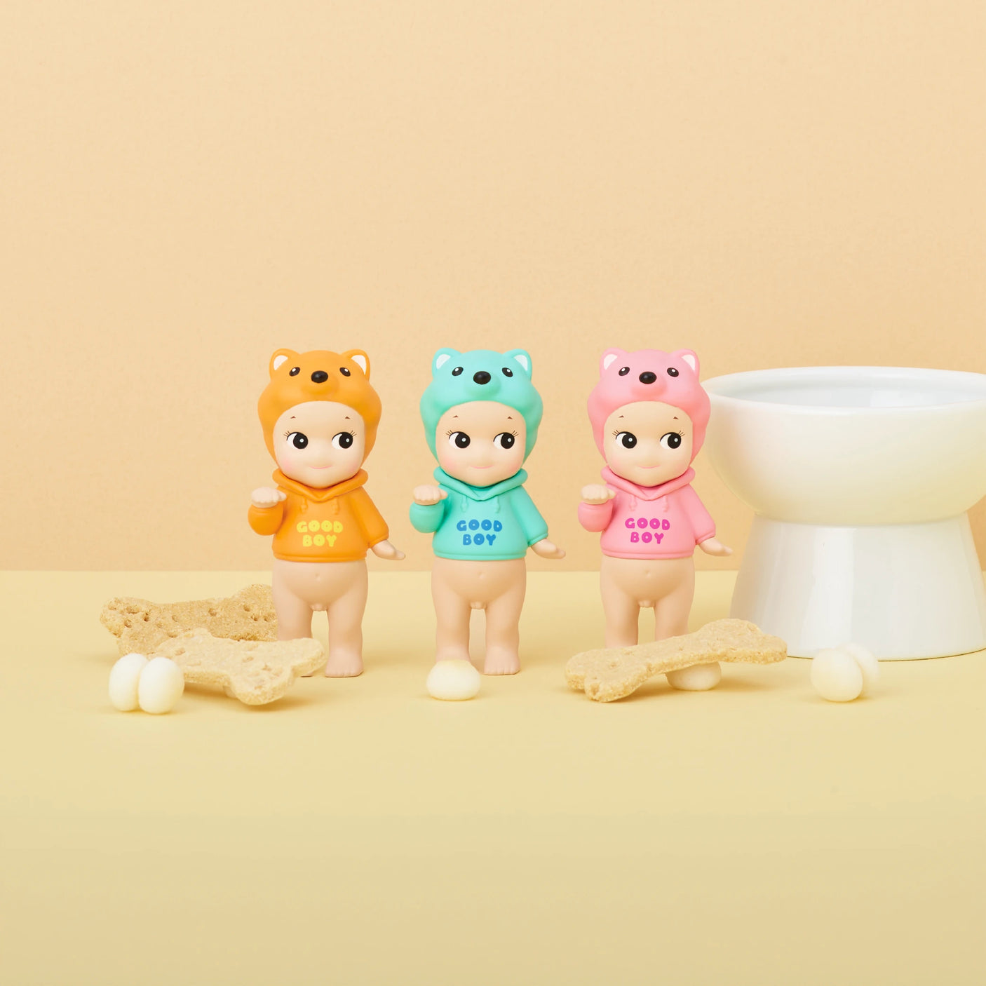 Three figures from the Sonny Angel Dog Time Series by Sonny Angel, each featuring animal-themed hats, are arranged in a row. These collectible mystery figures are set against a beige background, adorned with bone-shaped biscuits and small white balls.