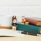 Two collectible figurines from the Sonny Angel Dog Time Series by Sonny Angel are positioned on books next to a mug and a pen. A charming "DOG FOOD" tin enhances the scene alongside these mystery figures.