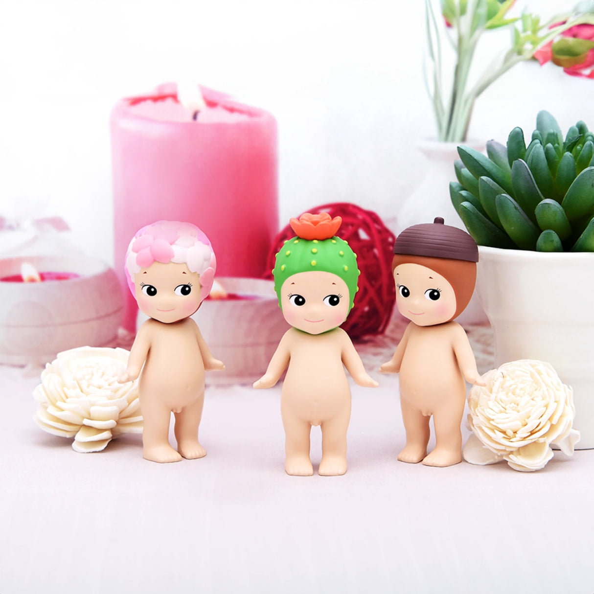 Three figures from the Sonny Angel Flower Series by Sonny Angel stand on a surface surrounded by candles and plants. Their plant-themed hats add a touch of charm, capturing the surprise and delight typical of a blind box experience.