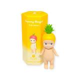 A small figurine wearing a pineapple hat is placed beside the yellow mystery box from the Sonny Angel Fruit Series Blind Box collection by Sonny Angel, featuring charming fruit-themed designs.