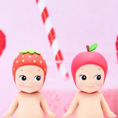 Two delightful figurines from the Sonny Angel Fruit Series Blind Box wear adorable strawberry and apple hats, set whimsically against a pink background, highlighting their playful fruit-themed design.