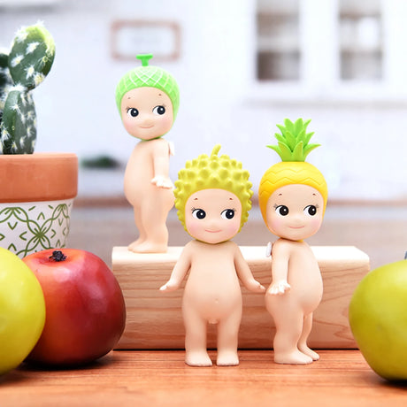 Three small figurines from the Sonny Angel Fruit Series Blind Box, each donning a fruit-themed hat—melon, durian, and pineapple—stand on a wooden block among a cactus and apples, capturing a whimsical fruit design. Could they become the latest addition to your mystery box?