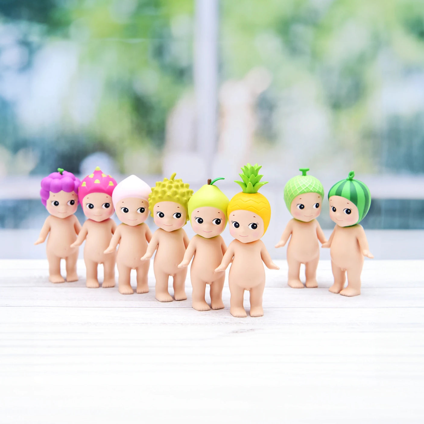 Eight small Sonny Angel figurines from the Fruit Series Blind Box, each wearing playful fruit-themed hats, stand lined up on a white surface, embodying the charm of their mystery box origin. The background is artistically blurred.