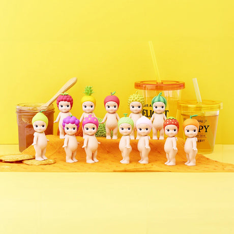 A group of Sonny Angel figurines from the Fruit Series, each adorned with fruit-themed headdresses, are displayed on a yellow surface with two jars and plastic cups in the background.