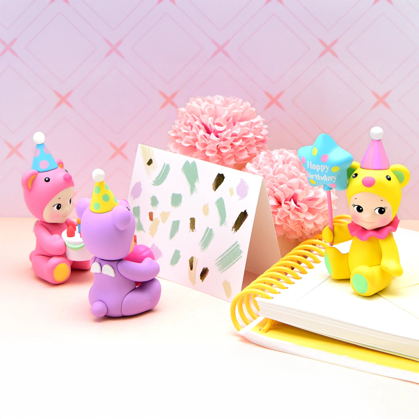 Three Sonny Angel Birthday Bear Gift figurines, adorned with party hats, are arranged near a card, pink flowers, and a notebook on a pastel background.