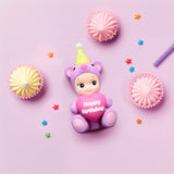 A charming Sonny Angel Birthday Bear Gift, featuring a toy dressed in a bear costume and party hat while holding a "Happy Birthday" heart, is surrounded by vibrant meringues and star-shaped confetti on a pink background. This delightful product from Sonny Angel makes the perfect gift for any celebration.