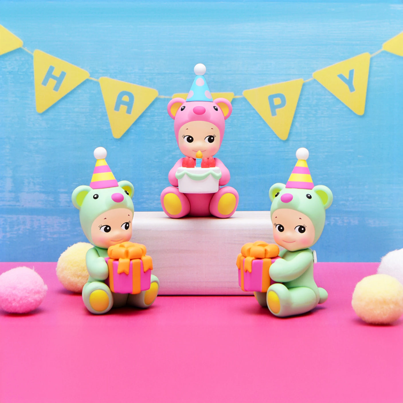 Three Sonny Angel toy figures sporting party hats, featuring a Birthday Bear Gift and two others, are seated on a pink surface with two holding presents beneath a backdrop displaying a "HAPPY" banner.