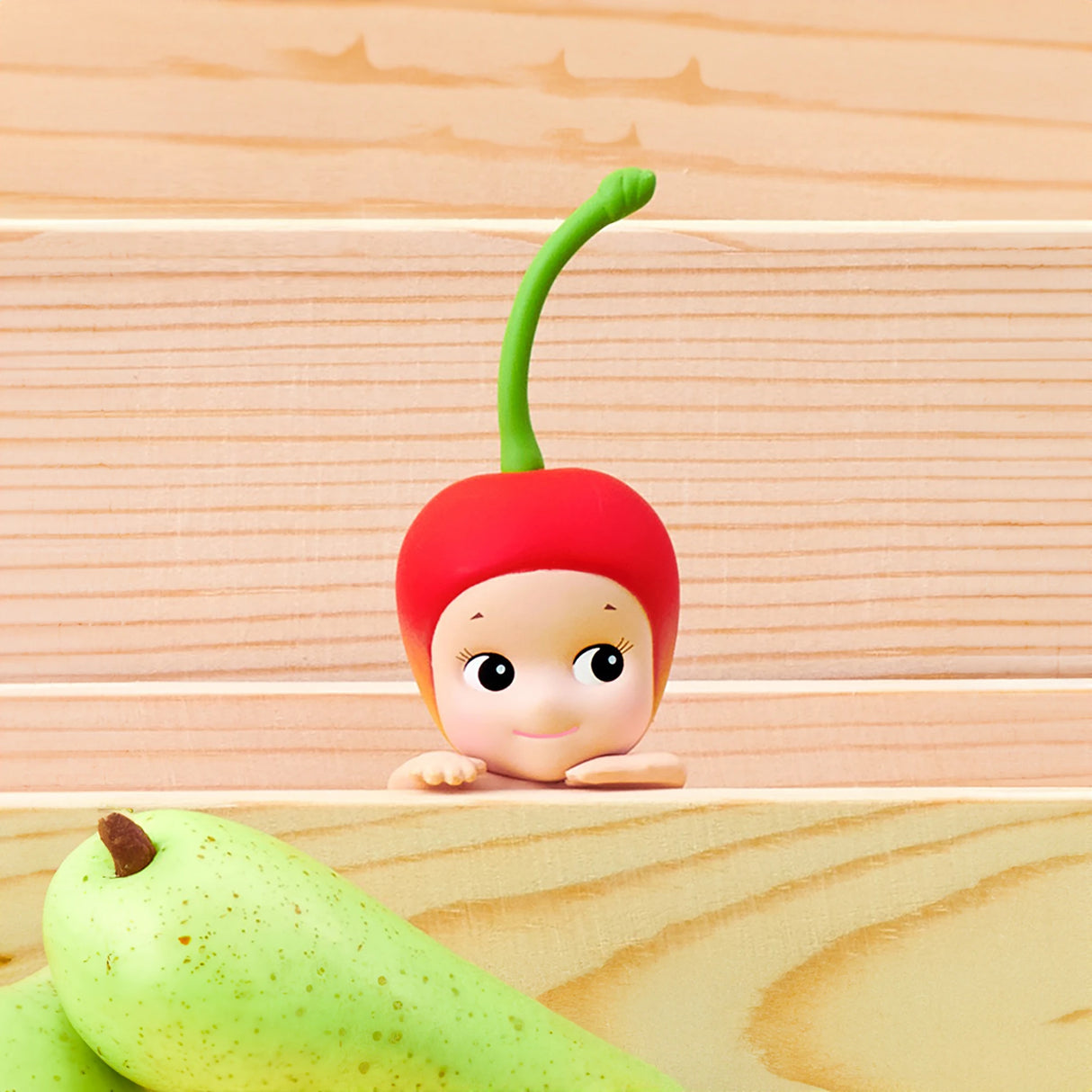 A Sonny Angel Hippers Blind Box - Harvest figure featuring a cherry-like red head and a playful facial expression peers over a wooden surface beside two ripe pears.