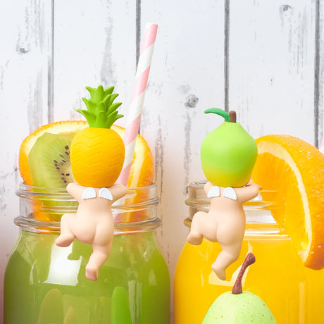 Two jars brimming with vibrant juice—one green and one yellow. Adorning the rims, Sonny Angel collectible figures from the Harvest series flaunt fruit-themed heads, resembling pineapple and apple. The cheerful scene is completed with slices of orange and kiwi garnishing the drinks, showcasing the delightful Sonny Angel Hippers Blind Box - Harvest collection.