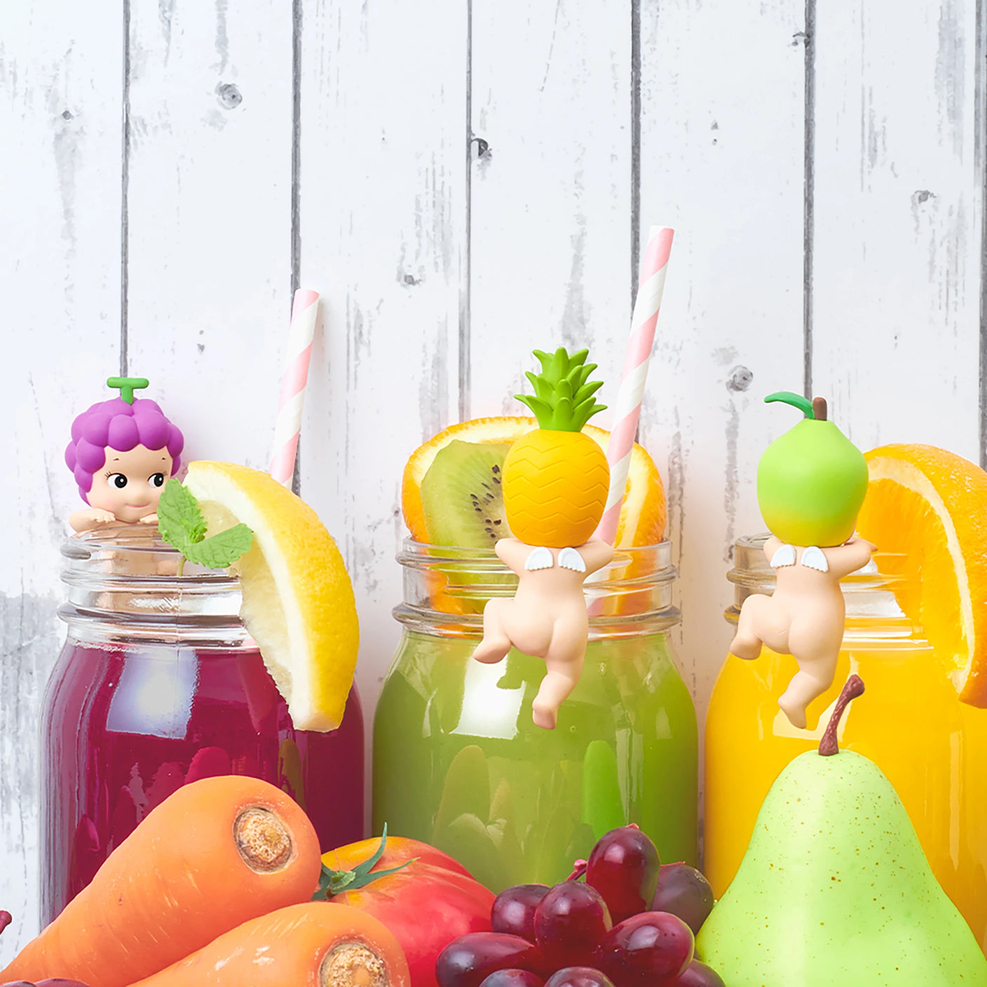 Brightly colored beverages in jars adorned with fruit garnishes and charming collectible figures from the Sonny Angel Hippers Blind Box - Harvest series. Surrounding the jars on a white wooden background are fruits such as grapes, pear, carrot, and pineapple.