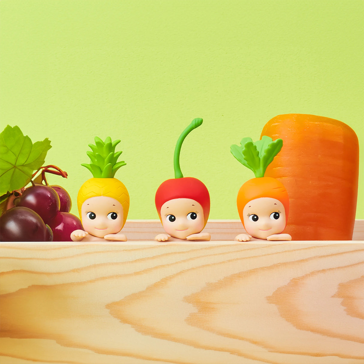 The Sonny Angel Hippers Blind Box - Harvest from the brand Sonny Angel boasts enchanting fruit and vegetable-themed figures, including a pineapple, radish, and carrot. These charming collectibles are beautifully arranged on a wooden surface with bunches of grapes and a large carrot in the background.