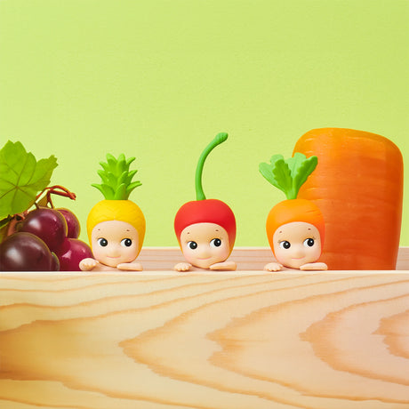 The Sonny Angel Hippers Blind Box - Harvest from the brand Sonny Angel boasts enchanting fruit and vegetable-themed figures, including a pineapple, radish, and carrot. These charming collectibles are beautifully arranged on a wooden surface with bunches of grapes and a large carrot in the background.