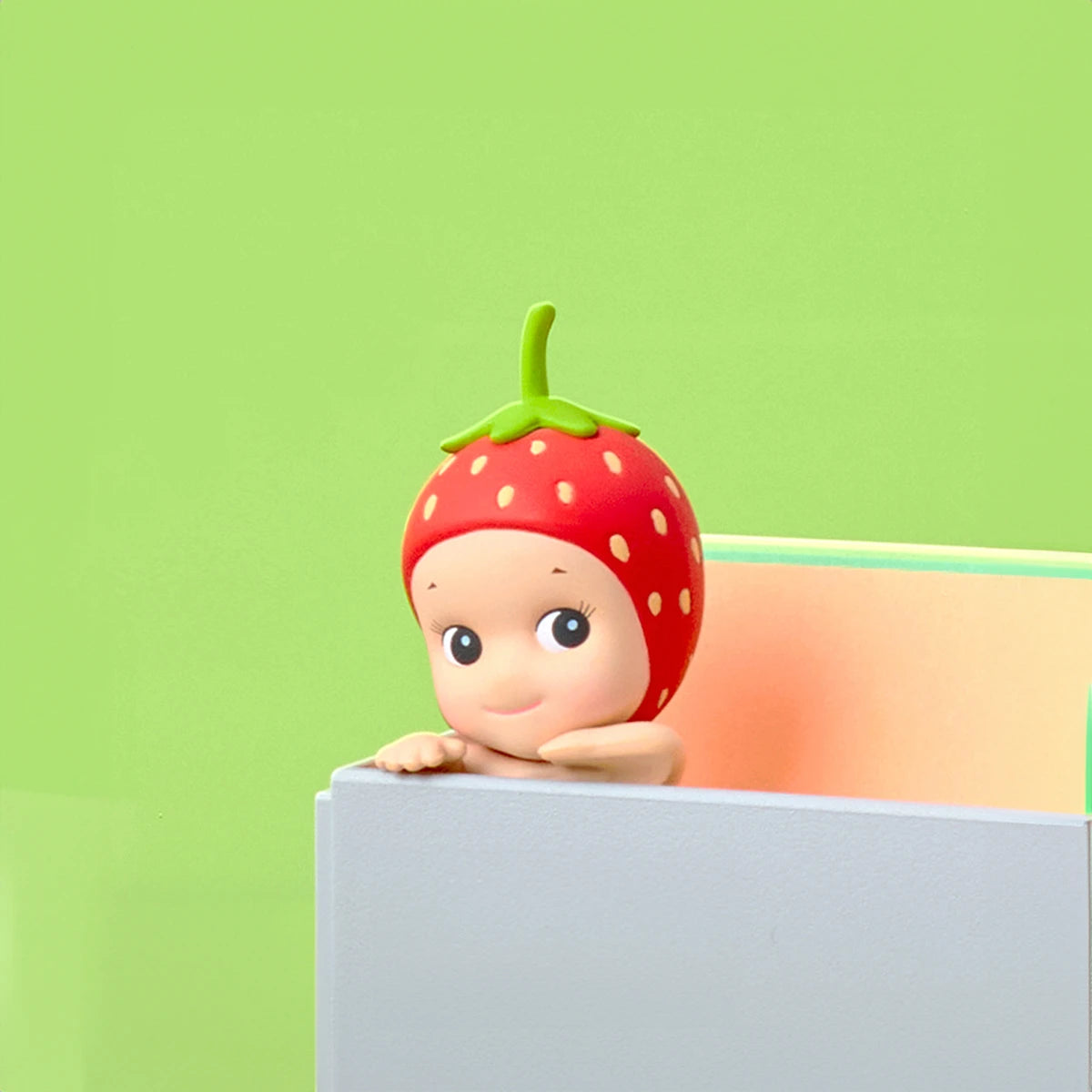 A Sonny Angel Hippers Blind Box - Harvest figure, by Sonny Angel, featuring a charming strawberry hat, peeks from behind a gray and pastel-toned panel set against a bright green background. This adorable figure is an excellent choice for enthusiasts of collectible toys.