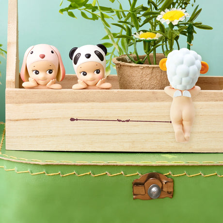 Three figures from the Sonny Angel Hippers Blind Box - Animals series, each adorned with a hat featuring a dog, panda, or sheep design, are placed near a wooden box and potted plant on a green surface. These charming figurines from Sonny Angel can be positioned using reusable adhesive for added convenience.