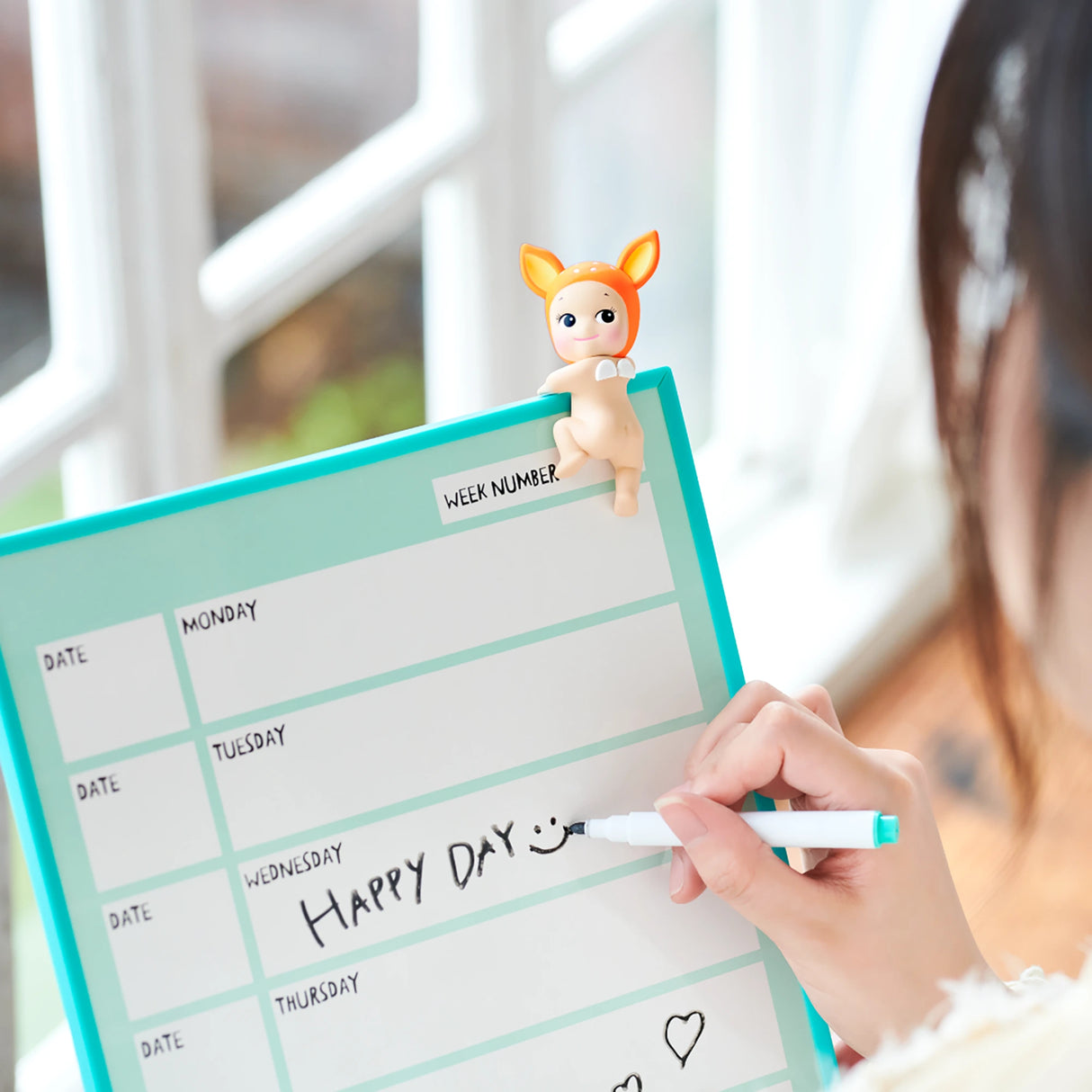 A person writes "Happy Day" with a smiley face on a weekly planner while a Sonny Angel Hippers Blind Box figure from the Looking Back series, one of those charming collectibles by Sonny Angel, perches playfully atop the board.