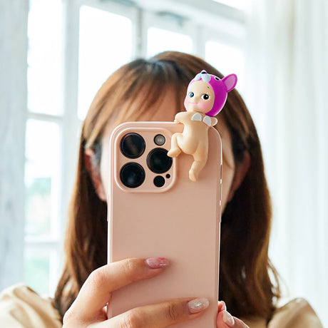 A person is holding a smartphone adorned with a Sonny Angel Hippers Blind Box - Looking Back figure from the Sonny Angel line. The doll perches atop, adding a whimsical touch to the tech-savvy scene.