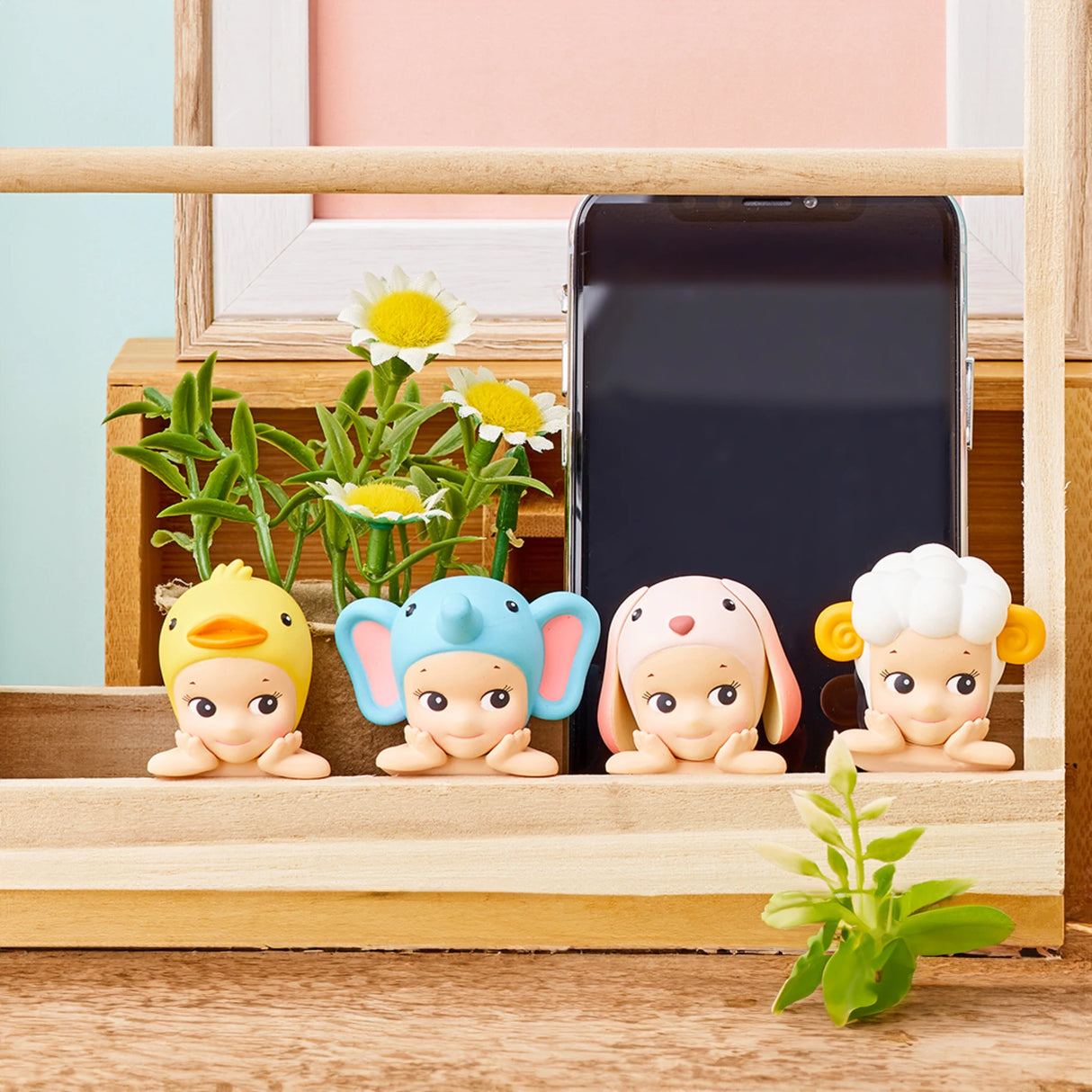 Four Sonny Angel figurines from the Hippers Blind Box - Animals series are lined up in front of a smartphone. Each figure displays an adorable animal hat, and they attach easily using reusable adhesive. The background features plants, adding to the charm of these pieces from the Sonny Angel brand.