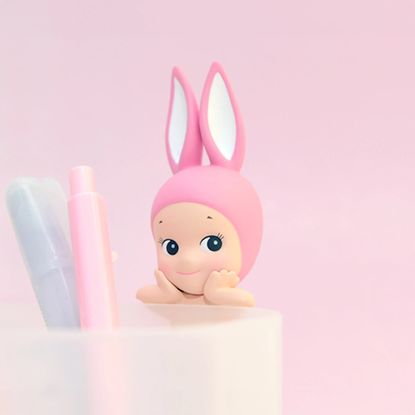 A charming Sonny Angel Hippers figure from the Animal Series, adorned with pink bunny ears and a smiling face, is showcased next to two markers in a white container against a pink background.