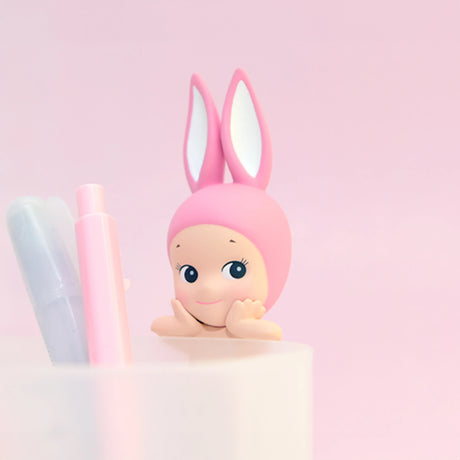 A charming Sonny Angel Hippers figure from the Animal Series, adorned with pink bunny ears and a smiling face, is showcased next to two markers in a white container against a pink background.