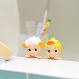 Two small, cartoonish figurines from the Sonny Angel Hippers Blind Box - Animals collection rest on a white surface. One figurine resembles a sheep with curly horns, while the other looks like a duck wearing a yellow beak and hat. In the background, blurred pens are held upright with reusable adhesive.