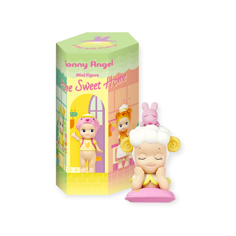 A small figurine with curly white hair, yellow horns, and a pink outfit sits cross-legged. Behind it is a decorative box labeled "Sonny Angel Home Sweet Home Series" from the Sonny Angel brand, inviting cozy companions to join the scene. Be on the lookout for Robby's cheeky cameo in this charming setting.