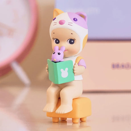 A charming figurine from the Sonny Angel Home Sweet Home Series by Sonny Angel features a doll dressed in a cat-themed outfit, seated on a stool with a green book and Robby's playful cameo, perfect for creating cozy companionship.