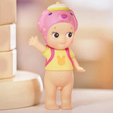 The Sonny Angel Home Sweet Home Series figure from the Sonny Angel brand features a charming toy character with a smiling face, wearing a pink animal hat and a yellow shirt adorned with a bear design, while carrying a pink backpack and standing on a smooth surface.