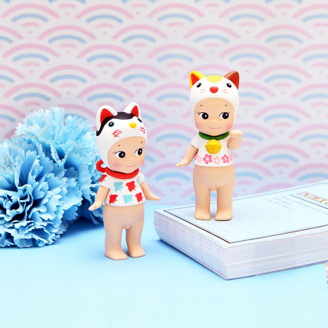 Two Sonny Angel Japanese Good Luck figurines stand on a notepad, each wearing a cat-themed hat. A blue flower sits beside them, while the background displays a rainbow pattern, adding an aura of good fortune to the scene.