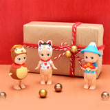 Three figures of Sonny Angel Japanese Good Luck toys, dressed in animal and mountain-themed costumes, are positioned before a gift box wrapped with a red ribbon. They are surrounded by gold spheres on a vibrant red background, capturing the essence of Japanese Lucky Charms.