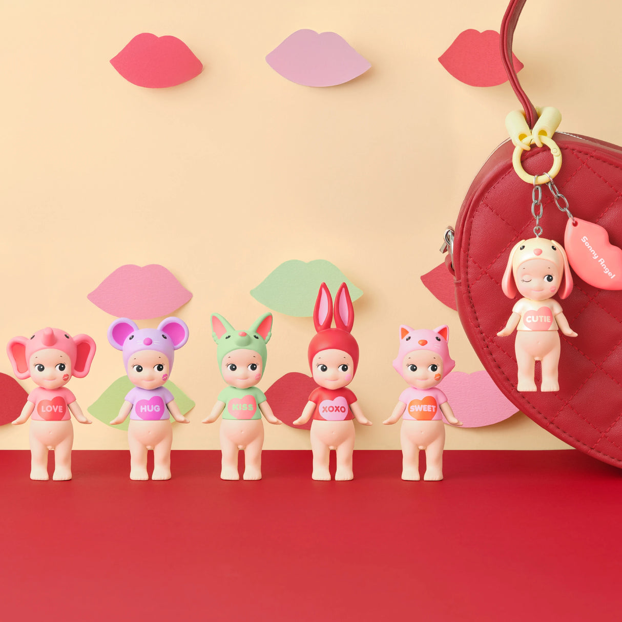 Five Sonny Angel figurines from the Kiss Kiss Series stand on a red surface, each with unique messages on their shirts. A red bag with a keychain lies to the right, surrounded by lips cut-outs for a playful vibe. This is part of the Sonny Angel Kiss Kiss Series - Blind Box collection.