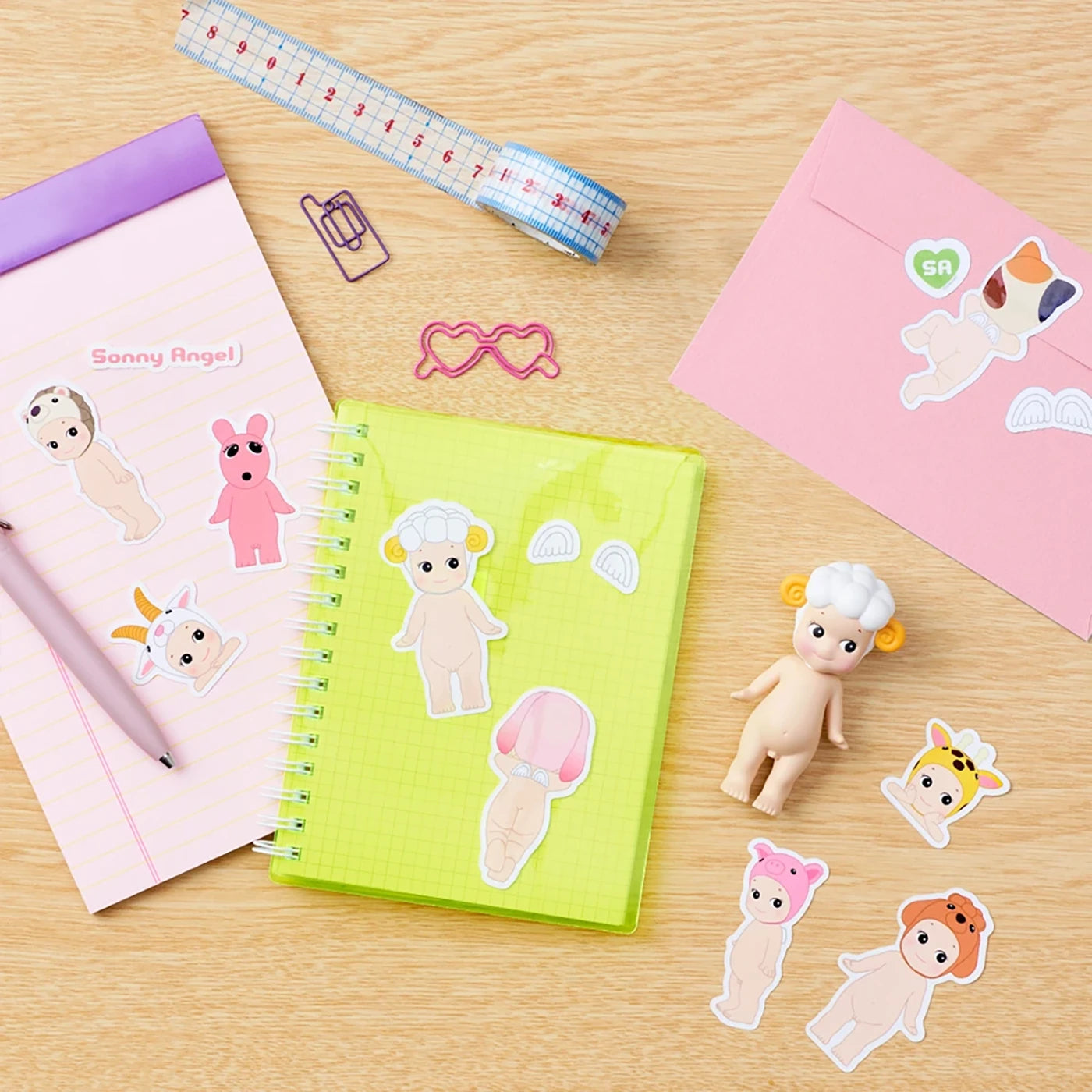 A desk with a yellow notebook, a pink notepad, a Sonny Angel Blind Box - Sticker Pack Series 1 featuring small figurines, paperclips, a measuring tape, and a pink envelope.