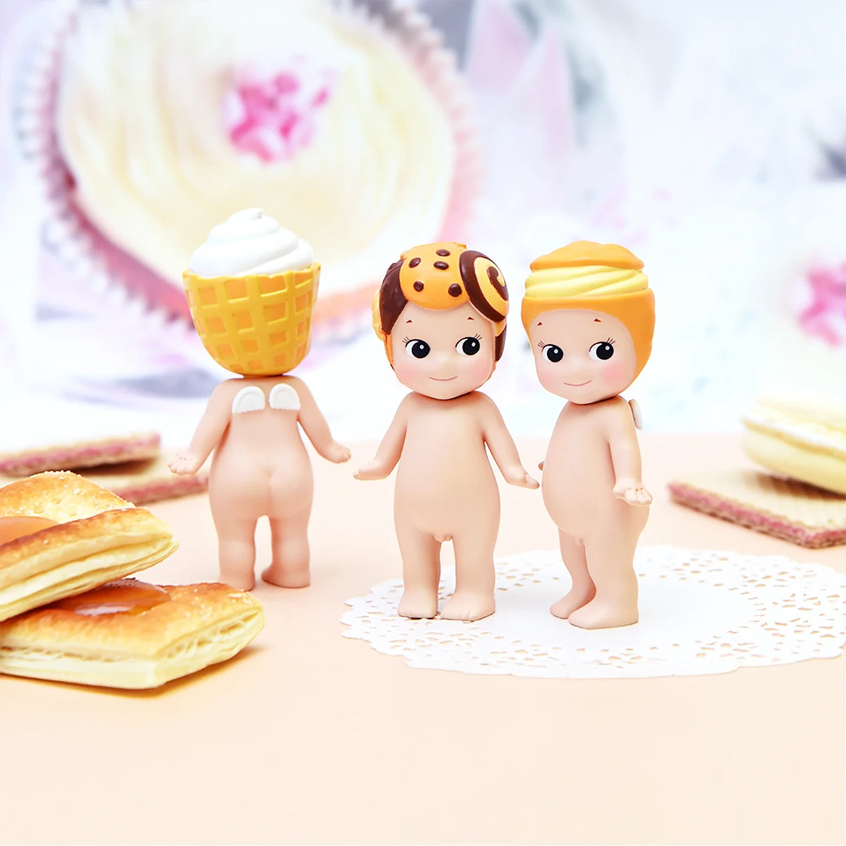 Three figures from the Sonny Angel Sweets Series Blind Box by Sonny Angel, each wearing a dessert-themed hat, are placed on a doily amidst an assortment of delicious pastries.