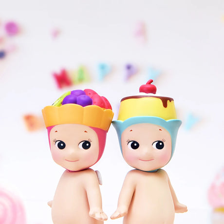 Two figures from the Sonny Angel Sweets Series Blind Box, each adorned with fruit and dessert-themed hats, stand side by side. They're set against a blurred background with colorful letters spelling "HAPPY," capturing the delightful mystery associated with Sonny Angel's collectible surprises.