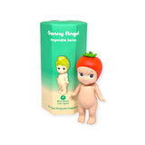 A cute figurine with a tomato hat stands proudly next to its packaging from the "Sonny Angel Vegetable Series Blind Box," part of the adorable lineup by Sonny Angel.