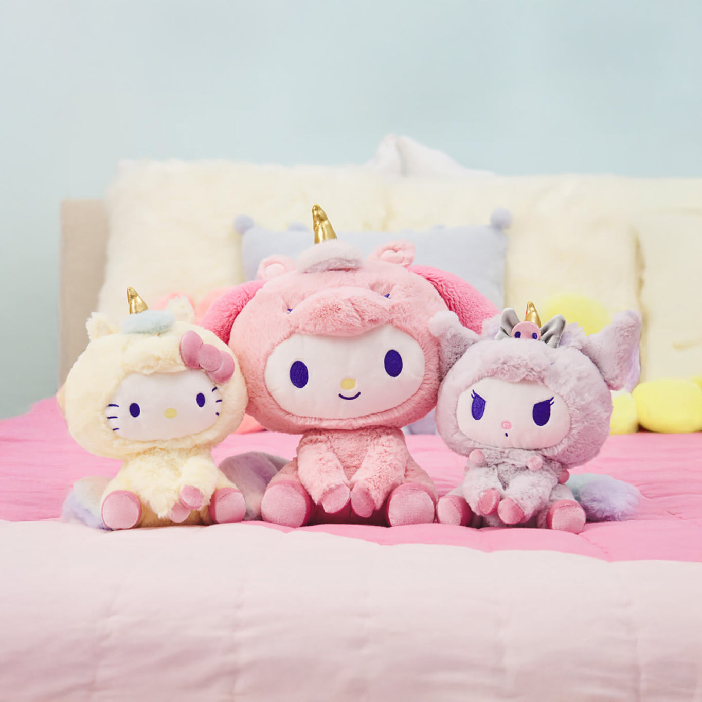 Three plush toys with unicorn horns rest on a pink bed, delighting Sanrio fans with a Hello Kitty-themed collection featuring a yellow Hello Kitty unicorn, a pink Cinnamoroll, and a gray My Melody.