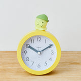 A yellow analog clock, adorned with a glow-in-the-dark Smiski Hippers figurine from the Smiski brand, featuring a green cap, is charmingly displayed on a wooden surface.