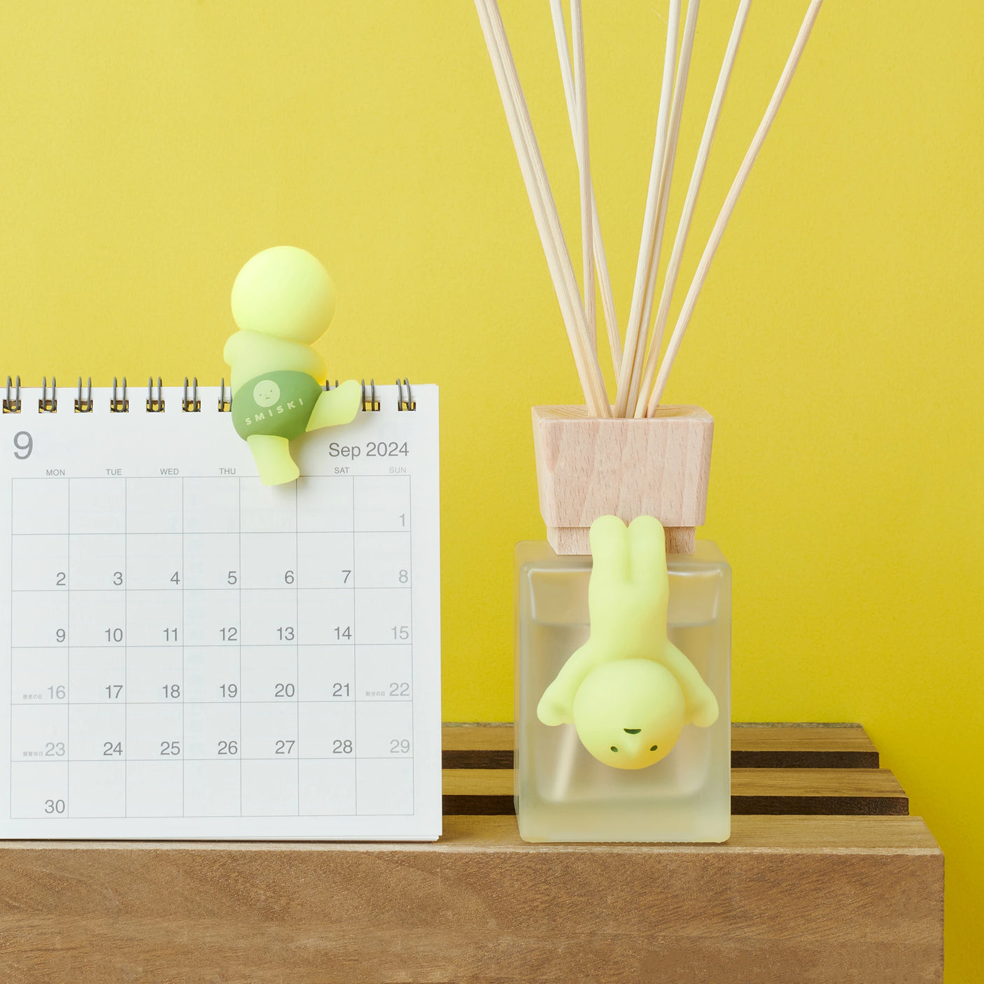 A small figurine from the Smiski Hippers Blind Box by Smiski is perched on a September 2024 calendar. Another similar glow-in-the-dark figurine hangs upside down playfully in a diffuser bottle, all set against a vibrant yellow background.