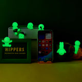 A collection of green glow-in-the-dark mini figures is artistically positioned around a smartphone, a mug, and a box against a dimly lit backdrop. A sign displays "Smiski Hippers Blind Box," showcasing the brand Smiski.