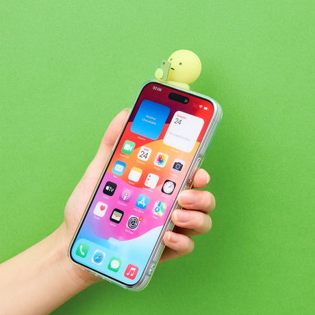 A hand holds a smartphone displaying a vibrant, app-filled screen against a green background. Perched at the top of the phone is a glow-in-the-dark character from the Smiski Hippers Blind Box by Smiski.