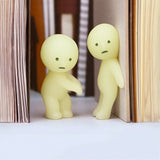 Two small, glowing figures from the Smiski Series 1 - Blind Box by Smiski are tucked between the open pages of books, their pale yellow round heads appearing to interact with each other in the dark.