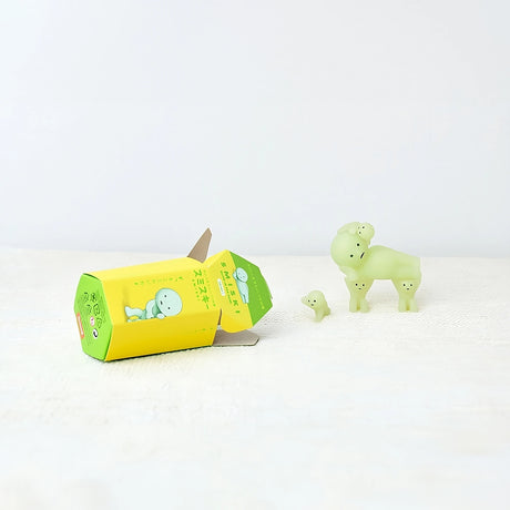 Two small, dog-like figurines from the Smiski brand's Series 1 sit next to an opened Smiski Series 1 - Blind Box on a white surface, ready to glow in the dark with their green hue.