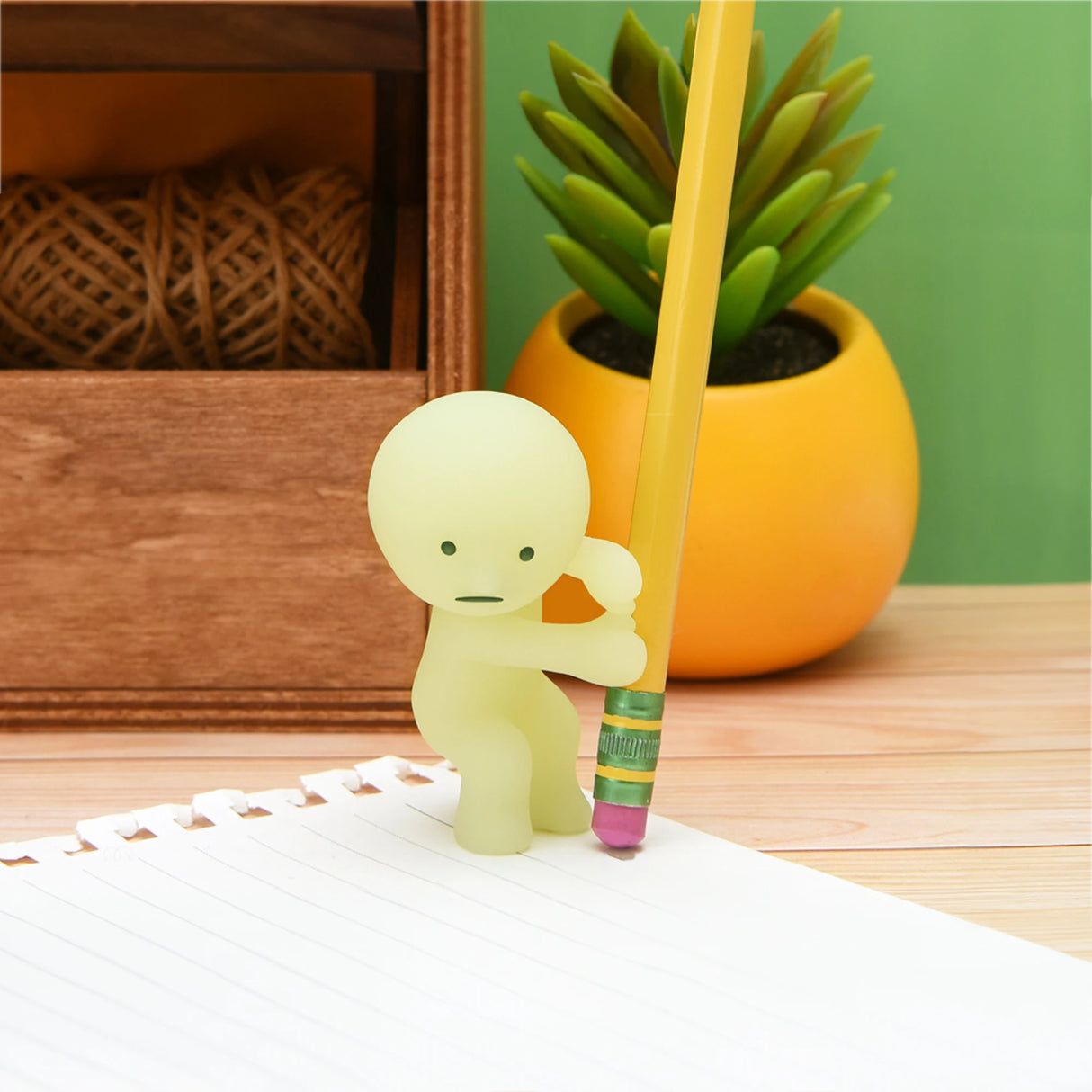 A small figurine from the Smiski Series 1 - Blind Box by Smiski holds a yellow pencil upright on a lined sheet of paper. In the background, a wooden box and a yellow pot with a green plant add charm to this whimsical scene. This glow-in-the-dark collectible is sure to delight fans of surprise blind boxes.